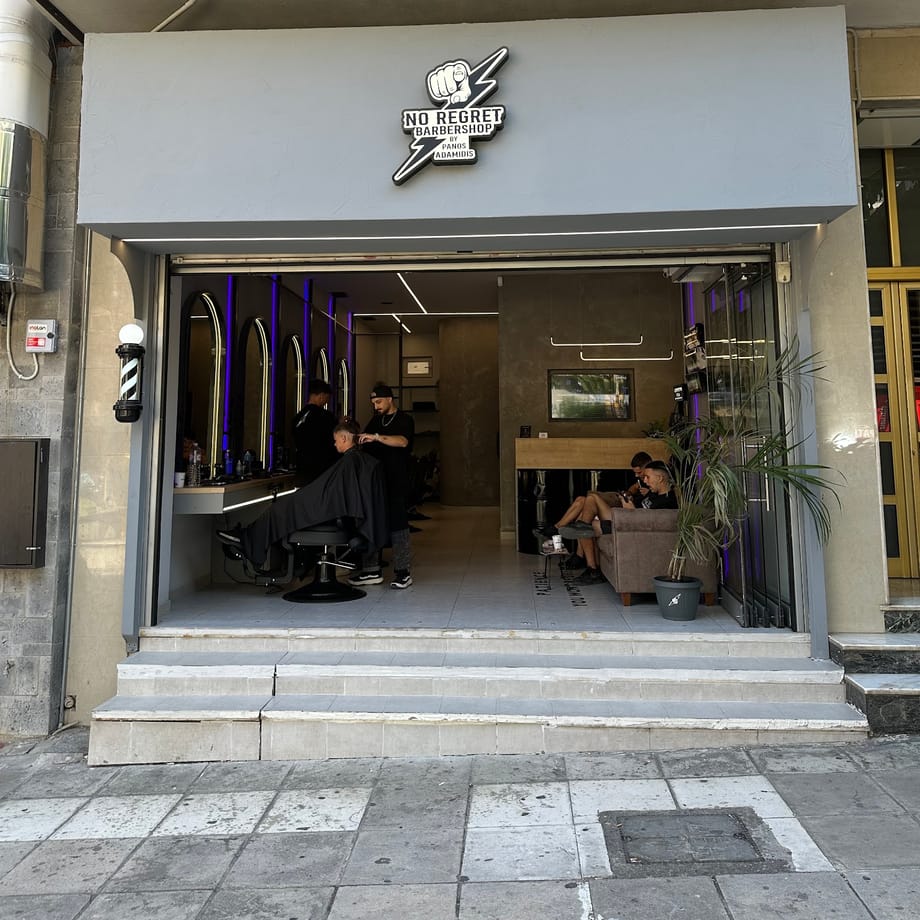 Η πρόσοψη του No Regret Barbershop στην Υμηττού 129: το ανάγλυφο σήμα με τη γροθιά, η μπαρμπερόμπαρα και η ανοιχτή είσοδος με τους μωβ φωτισμένους καθρέφτες